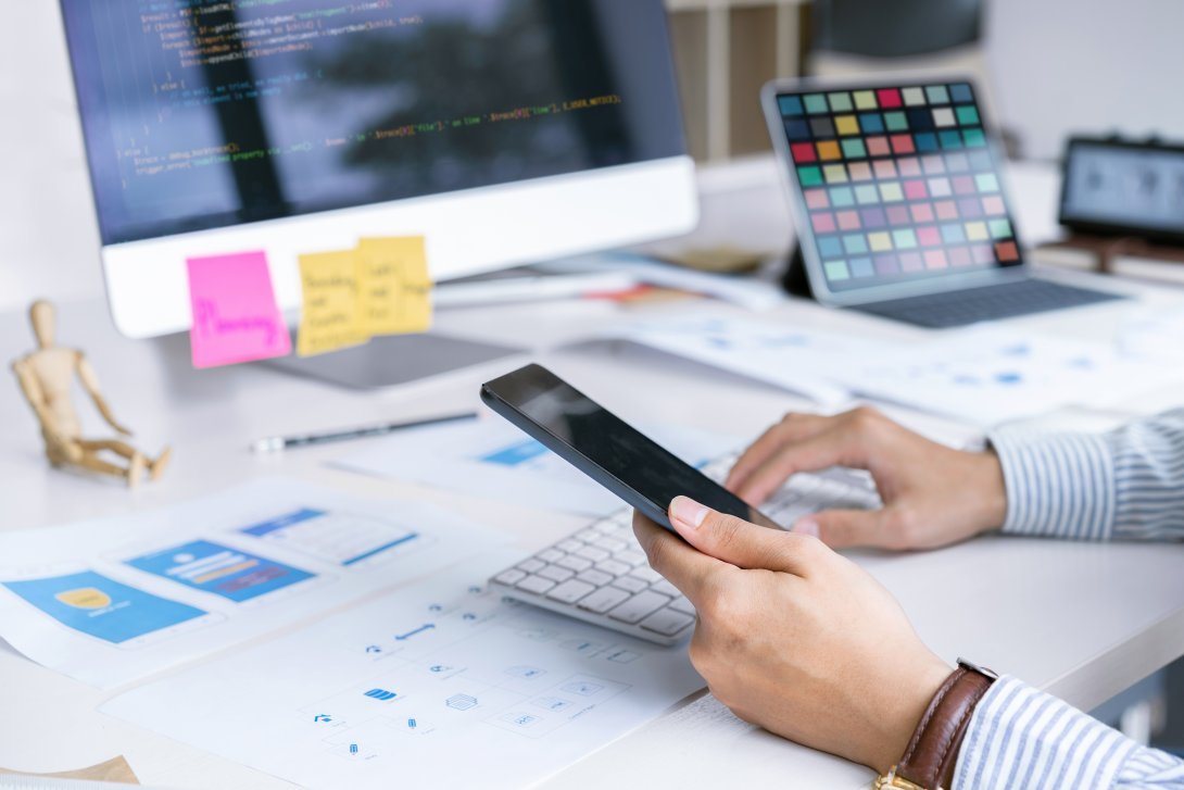 Scrivania con fogli, schermo di computer, tastiera e mani che consultano uno smartphone